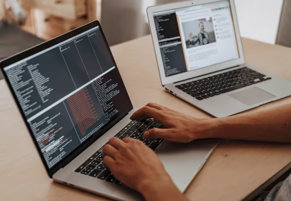 Deux personnes travaillant sur deux ordinateur. L'une des personne montre du doigt des lignes de code sur son ordinateur